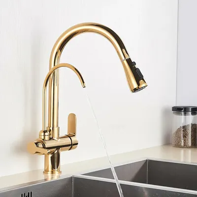 A gold kitchen faucet with a pull-out sprayer is shown in use, with water flowing into a sink. A jar of spices is visible in the background.