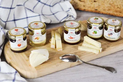 A set of five small jars of sauces and cheese on a wooden board, with a checkered cloth and bread in the background.