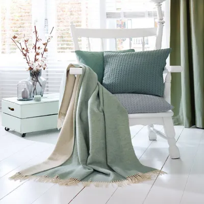 A cozy living room scene with a white chair draped with a green and cream plaid blanket, two decorative pillows, and a vase of flowers on a side table.
