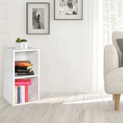 A white corner shelf unit with books and plants, placed in a room with a sofa and a window.