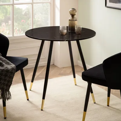 A round black marble-look dining table with black metal legs and gold accents, set in a room with two black chairs and a window in the background.