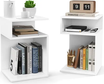 Two white S-shaped side tables are shown against a white background. The table on the left has books, a plant, and a teapot. The table on the right has books, pens, and a clock.