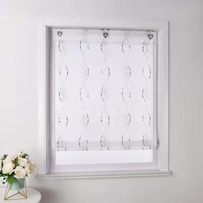 A white roller blind with a brown embroidered pattern is shown hanging in a window frame. A small bouquet of white flowers in a teal pot sits on a small table to the left of the window.