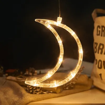 A crescent moon-shaped LED light decoration hangs against a dark background, with some decorative items like pinecones visible in the foreground.