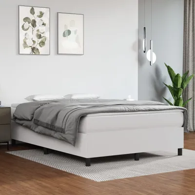 A white boxspring bed with a grey duvet and pillows, set in a minimalist bedroom with artwork on the wall and a plant in the corner.