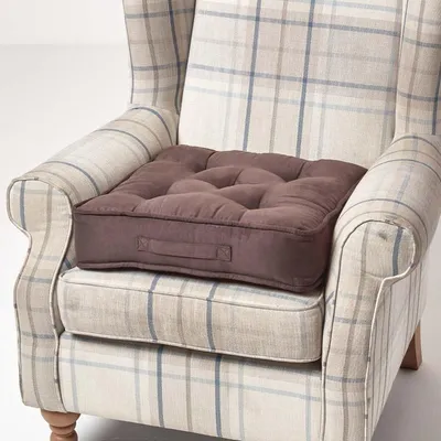 A brown tufted seat cushion is placed on a plaid armchair.