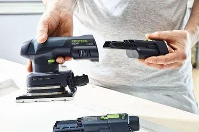 A person is holding a Festool Akku-Rutscher RTSC 400-Basic-ERGO sander and a battery pack, demonstrating how they connect.