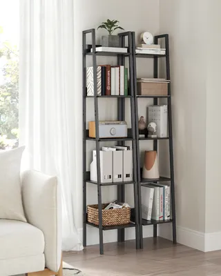 A narrow, multi-tiered ladder bookshelf filled with books and decorative items, placed next to a white sofa and in front of a window with sheer curtains.