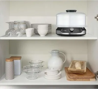 A kitchen cabinet shelf displaying various kitchenware, including a steamer, bowls, glasses, and a carafe.