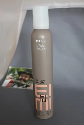 A bottle of Wella EIMI Natural Volume Styling Mousse is shown in the foreground, with a blurred image of a person in the background.