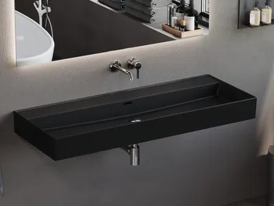 A modern black matte washbasin mounted on a gray wall, with a faucet and drain visible. The reflection in the mirror above shows a white bathtub and toiletries.