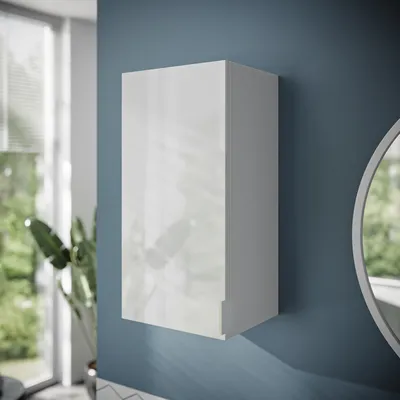 A white bathroom cabinet is mounted on a blue wall next to a round mirror. A large window with blinds and a potted plant are visible in the background.