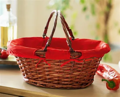 A woven basket with a red fabric liner sits on a wooden surface, with a bottle and peppers in the background.