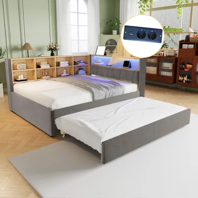 A bedroom scene featuring a grey upholstered bed with a pull-out trundle bed, a wooden bookshelf headboard with integrated LED lighting, and a close-up inset showing electrical outlets and USB ports.