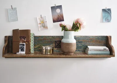 A rustic wooden shelf mounted on a white wall, decorated with a vase of flowers, books, a small metal container, and some postcards clipped to the wall.