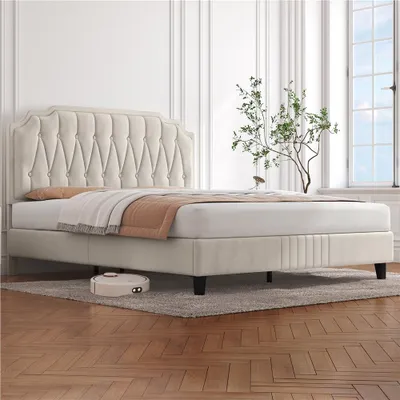 A beige upholstered bed with a tufted headboard is shown in a well-lit room with white paneled walls and a large window. A small robotic vacuum cleaner is visible on the rug beneath the bed.