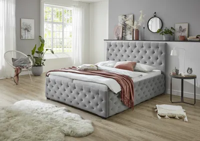 A bedroom scene with a tufted bed as the main focus, decorated with pillows and a throw blanket. A chair, plant, and decorative items are also visible.
