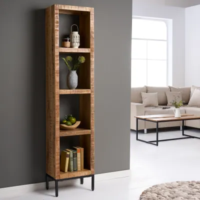 A tall, narrow wooden bookshelf with four shelves, filled with decorative items and books, stands against a gray wall in a living room setting. A sofa and coffee table are visible in the background.