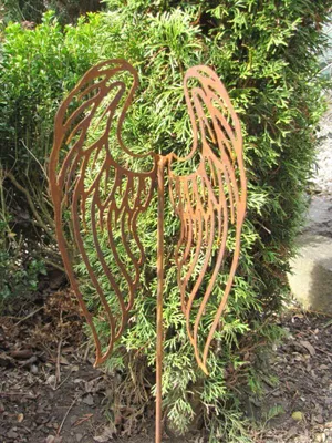 A rusted metal garden stake shaped like angel wings is planted in the ground in front of a green bush.