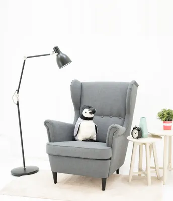 A plush penguin sits on a gray armchair next to a floor lamp and a small side table with decorative items.