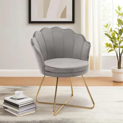 A gray velvet accent chair with gold metal legs is shown in a well-lit room with a plant and books in the foreground.