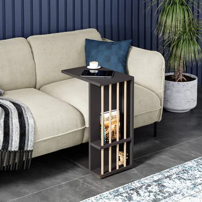 A dark gray side table with vertical wooden slats is placed next to a beige sofa in a living room setting. A tablet and a cup of coffee are on the table, and a book is in the lower shelf. A blanket is draped over the sofa, and a potted plant is visible in 