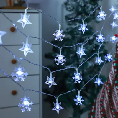 A string of LED lights shaped like stars and snowflakes, illuminated with a cool white light, is draped in front of a blurred Christmas tree and a white cabinet.