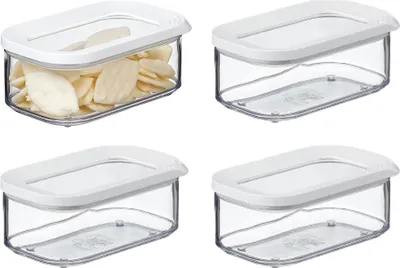 Four clear plastic food storage containers with white lids are arranged in a two-by-two grid against a white background. One container is filled with light-colored, flat food items, while the other three are empty.