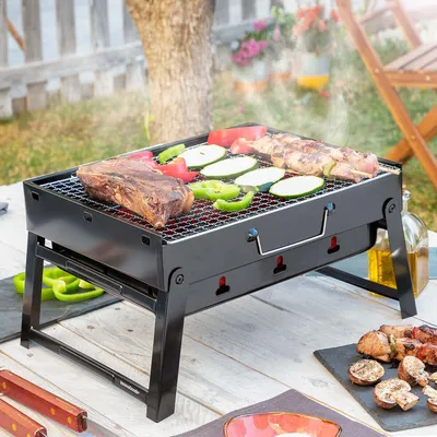 A portable charcoal grill with food cooking on it, set up on a table outdoors.