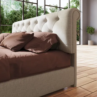Close-up side view of a tufted upholstered bed frame with brown bedding and pillows, set against a large window with greenery and a potted plant in a bright room with wooden floors.
