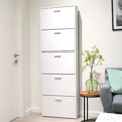 A white shoe cabinet with multiple drawers is shown in a room next to a door and a sofa.