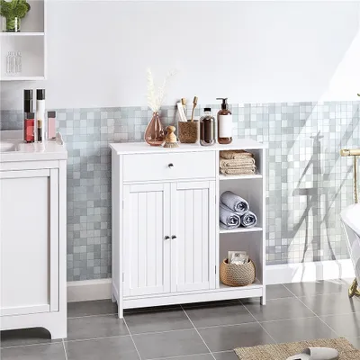 A white bathroom cabinet with a drawer and two doors, placed next to a vanity with bottles and a sink. The wall behind has a mosaic tile pattern.