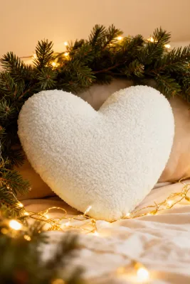 A white heart-shaped pillow is placed on a bed, surrounded by pine branches and fairy lights, suggesting a festive or romantic setting.