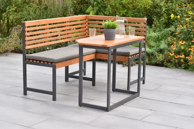 An outdoor seating set with a corner bench and a small table, placed on a patio with plants in the background.
