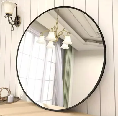 A round mirror with a black frame is displayed on a wooden surface against a white paneled wall. A wall sconce and a chandelier are reflected in the mirror.