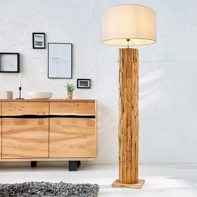 A wooden floor lamp with a linen shade stands next to a wooden sideboard in a room with a white textured wall.