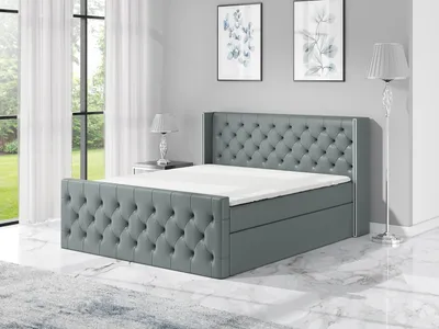 A grey upholstered boxspring bed with a tufted headboard and footboard, shown in a bedroom setting with two framed pictures on the wall and a floor lamp.