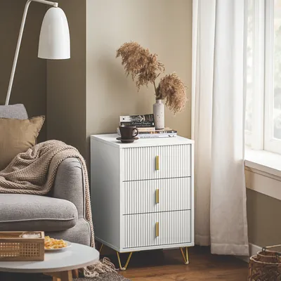 A modern living room scene featuring a white ribbed nightstand with gold legs, a sofa, a floor lamp, and a window with white curtains.