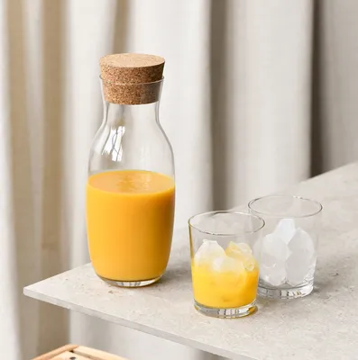 A glass carafe filled with orange juice and two glasses, one with orange juice and ice, the other with ice cubes, are placed on a countertop.