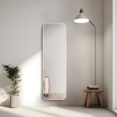 A full-length mirror is placed against a wall next to a potted plant and a floor lamp. A small wooden stool with folded towels sits beside the mirror.