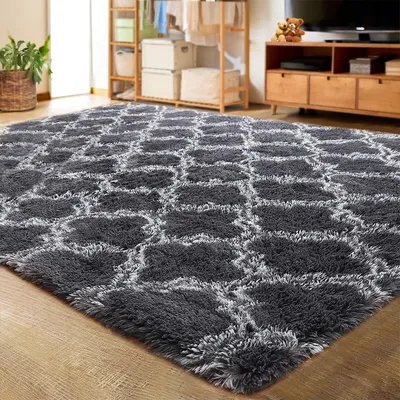 A fluffy gray rug with a geometric pattern is laid out on a wooden floor in a room. In the background, there is a wooden shelf unit with storage boxes and clothes, and a wooden TV stand with a television and decorative items.