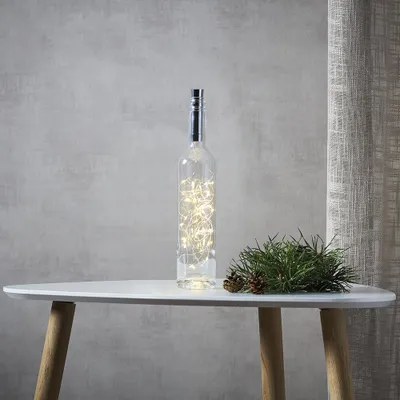 A glass bottle filled with warm white LED lights sits on a white table next to a sprig of pine and pinecones.