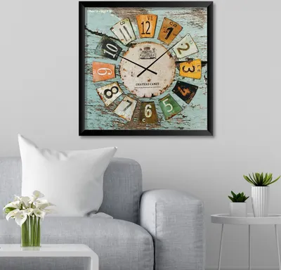 A wall clock with a vintage license plate design is displayed on a wall above a sofa. A white pillow and a vase with white flowers are on the sofa.