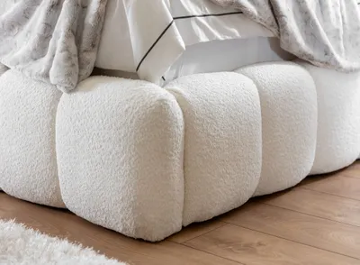 Close-up of a white textured bed skirt with a fluffy blanket and pillows on top, resting on a wooden floor.
