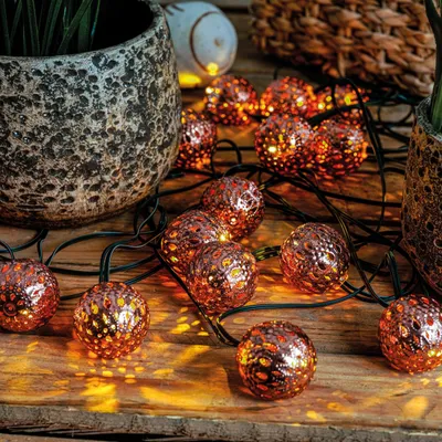 A string of copper-colored fairy lights with intricate patterns, illuminated and casting a warm glow on a wooden surface, next to a textured planter.
