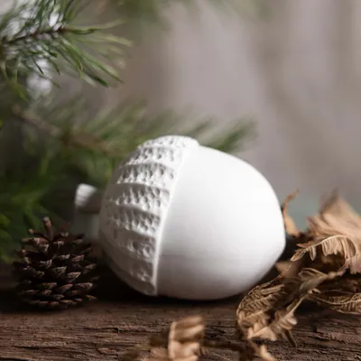 A white acorn-shaped ornament sits on a wooden surface, next to a pine cone and dried leaves, with a pine branch in the background.