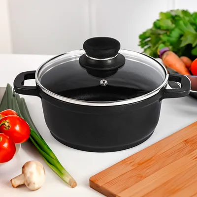 A black saucepan with a glass lid sits on a white countertop, surrounded by fresh vegetables and a wooden cutting board.