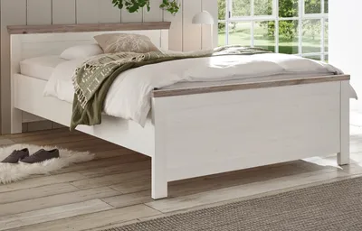 A bedroom scene with a bed, blanket, and pillows, set against a window with greenery outside.