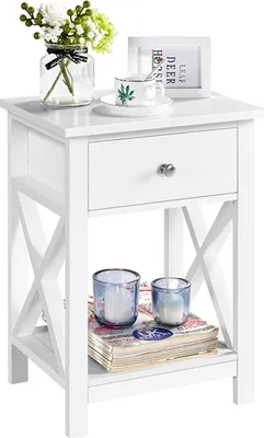 A white nightstand with a drawer and shelf, decorated with a vase of flowers, a teacup, a framed picture, and two candles on books.