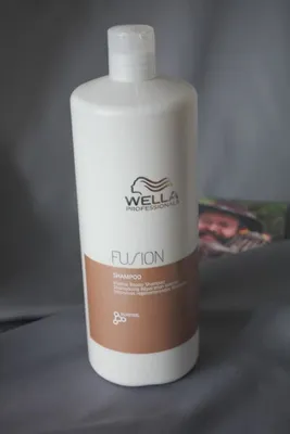 A bottle of Wella Professionals Fusion Intense Repair Shampoo is placed on a gray fabric background. A blurred image of a person wearing a hat is visible in the background.